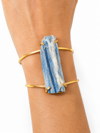 A close-up of a person's wrist wearing a gold cuff bracelet with a large rectangular blue stone centerpiece.