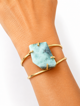 A close-up of a gold wrap cuff bracelet worn on a wrist, featuring a large, faceted light blue-green stone with natural dark veining.