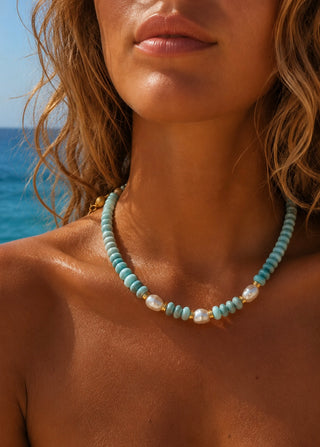 A close-up of a woman at the beach wearing a light blue and white beaded necklace with small gold-toned spacer beads.