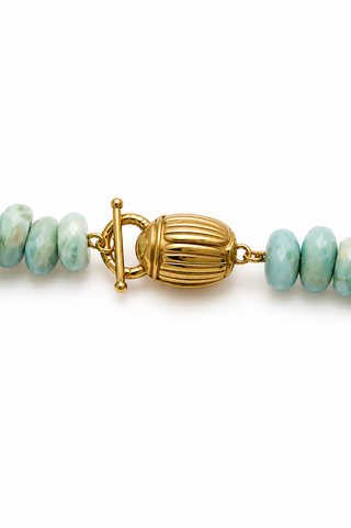 A close-up of a beaded necklace featuring light blue-green marbled beads and a prominent ribbed gold toggle clasp on a white background.