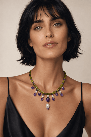 A woman with short dark hair wears a green beaded necklace with purple teardrop beads and a white pearl pendant, paired with a black satin top against a warm beige background.