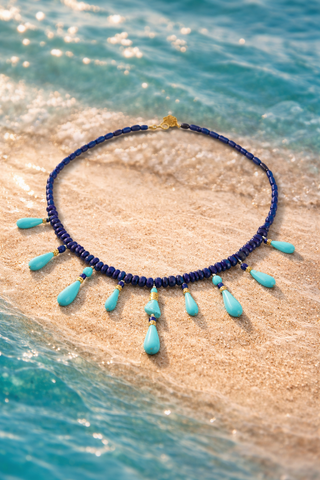 Navy blue beaded necklace with turquoise drops on a sandy beach with ocean waves.