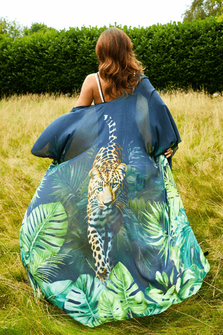 Person in a field wearing a navy and emerald kimono shawl with tropical leaf and leopard artwork, arms extended to show fabric movement.